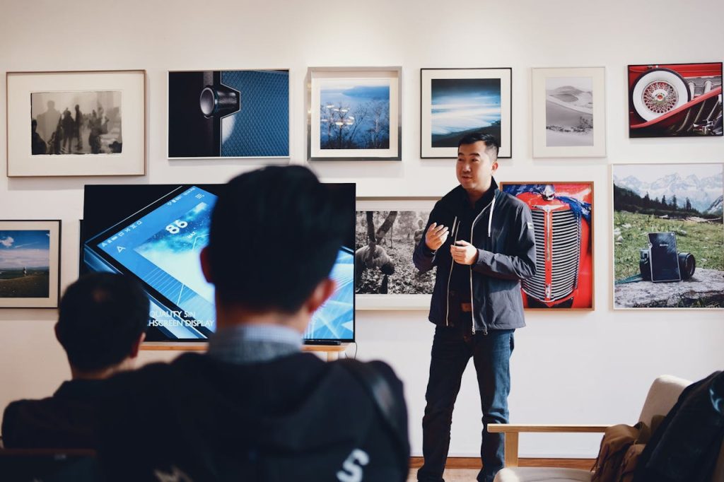 man-beside-flat-screen-television-with-photos-background-716276 Man giving a presentation in a modern art gallery setting, engaging audience.